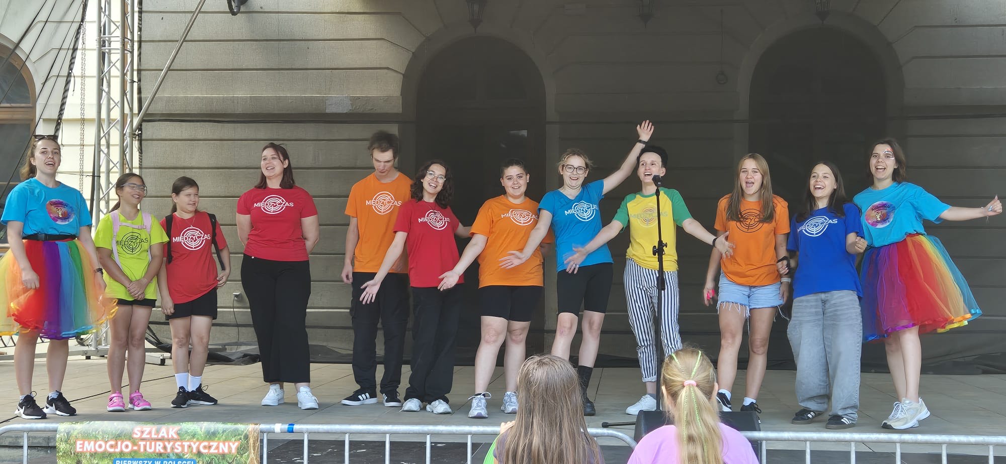 Dzień. Na scenie grupa osób ubranych w kolorowe podkoszulki