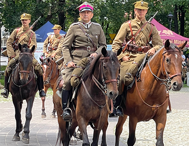Park, dzień. Aleja z kostki. Ułani w mundurach jada na koniach