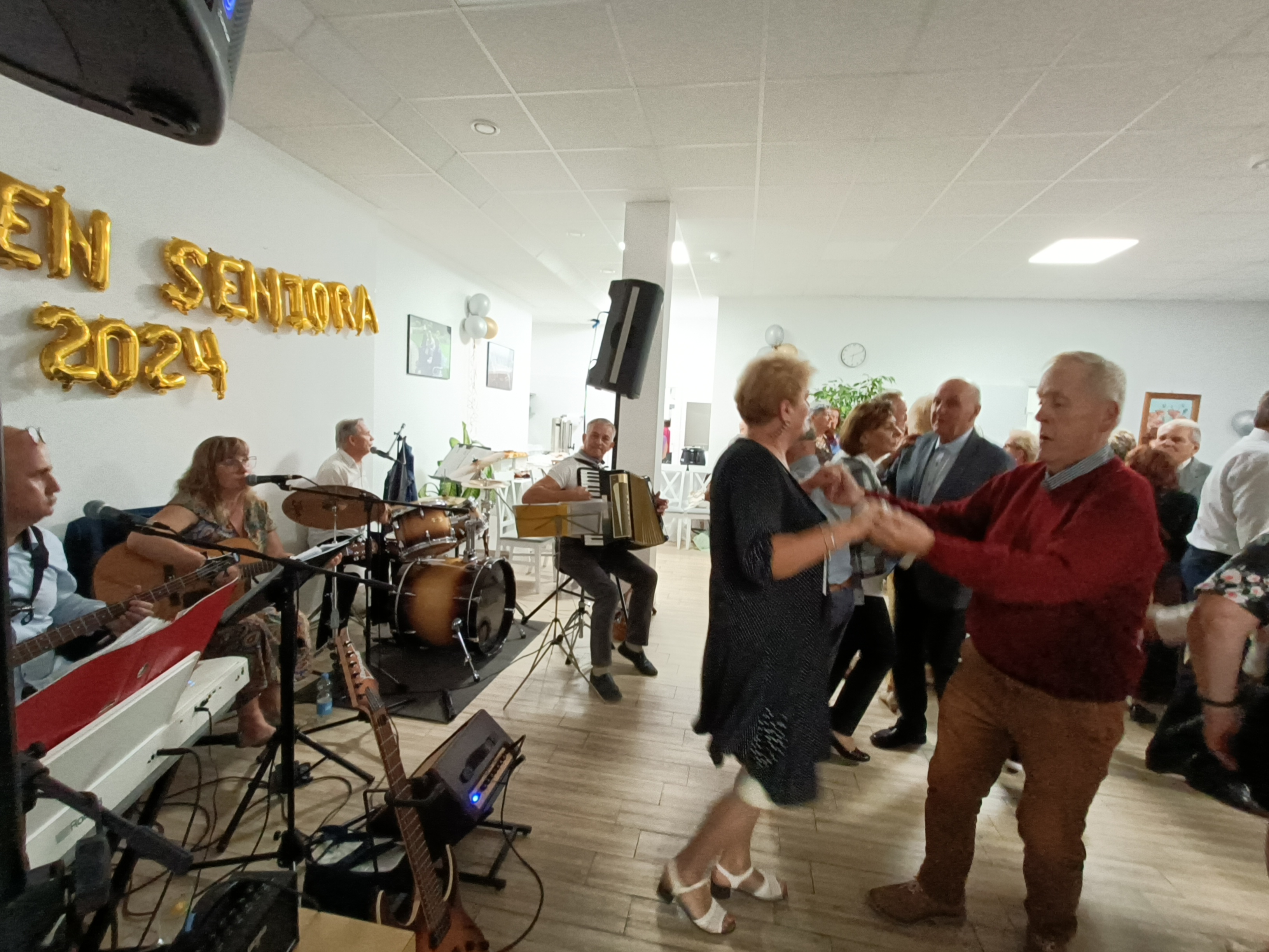 Pomieszczenie. Seniorzy tańczą. Po lewej gra orkiestra. Na jasnej ścianie z balonów ułożony napis dzień seniora 2024