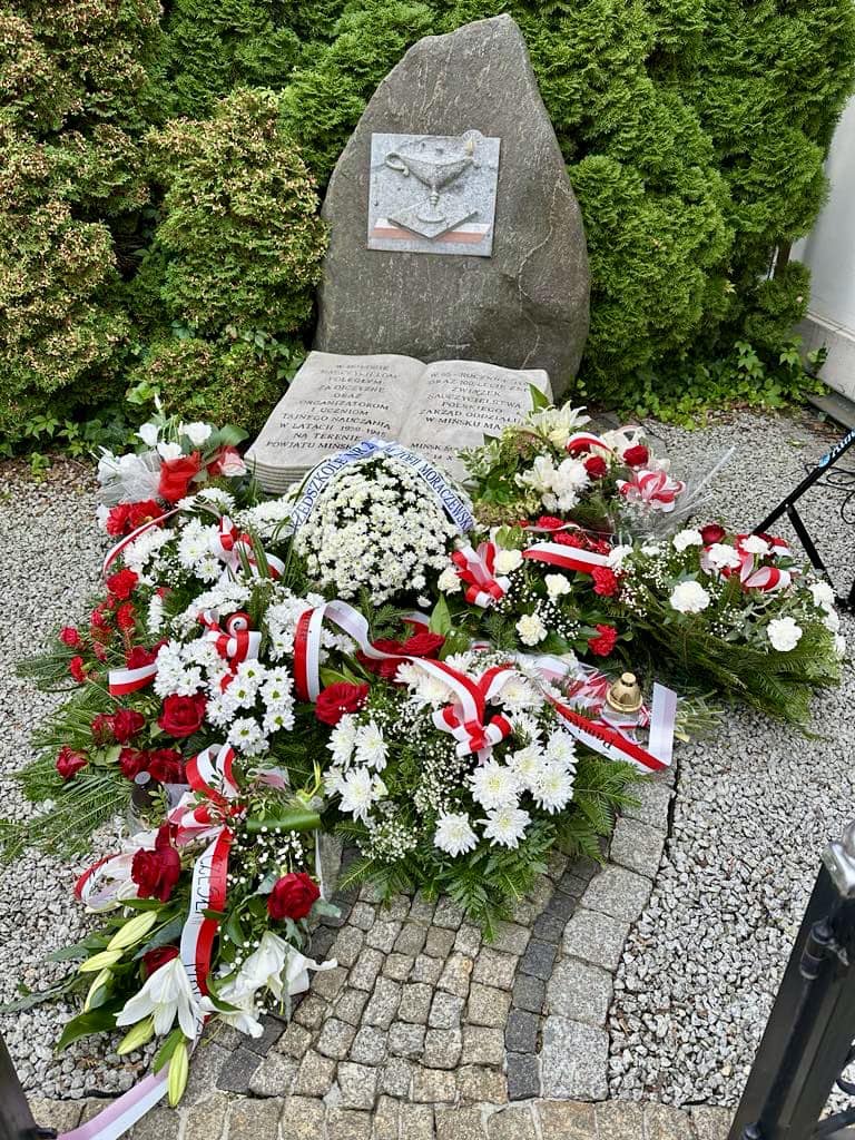Pomnik z kamienia, przed nim kamienna księga. Na przodzie wiązanki z biało-czerwonych kwiatów i szarf. Naokoło małe kamyczki i w tle zielone krzewy