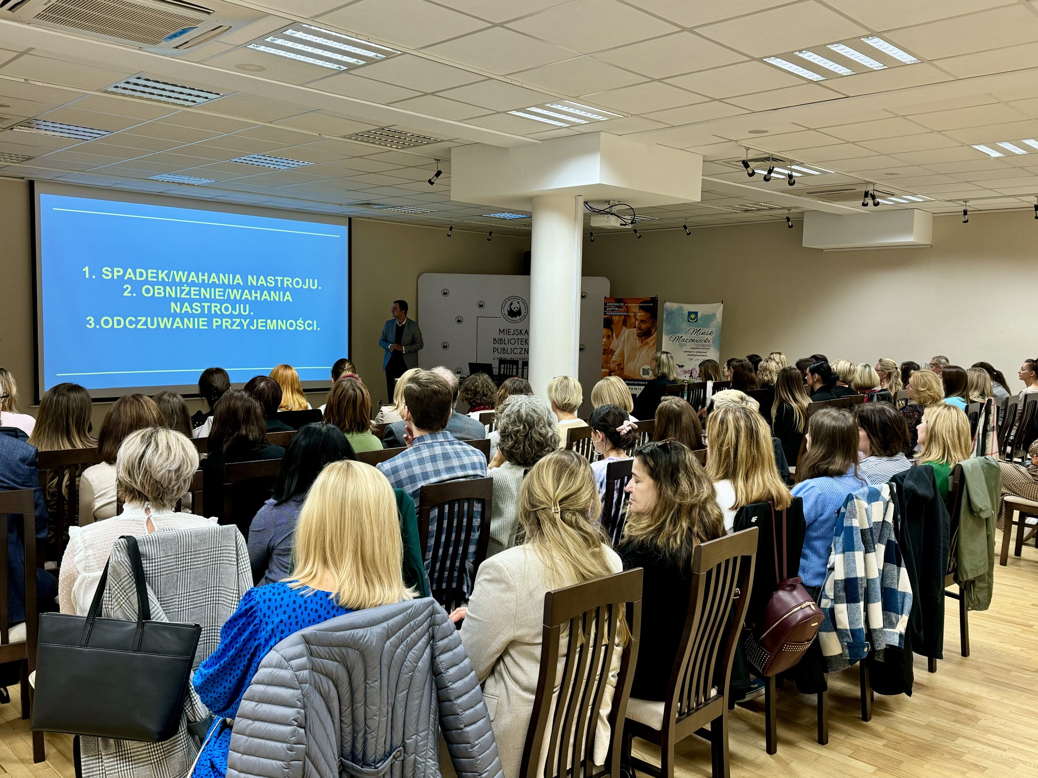 Sala konferencyjna. Na krzesłach siedzą osoby. Na przodzie mężczyzna opowiada stojąc przy dużym niebieksim ekranie