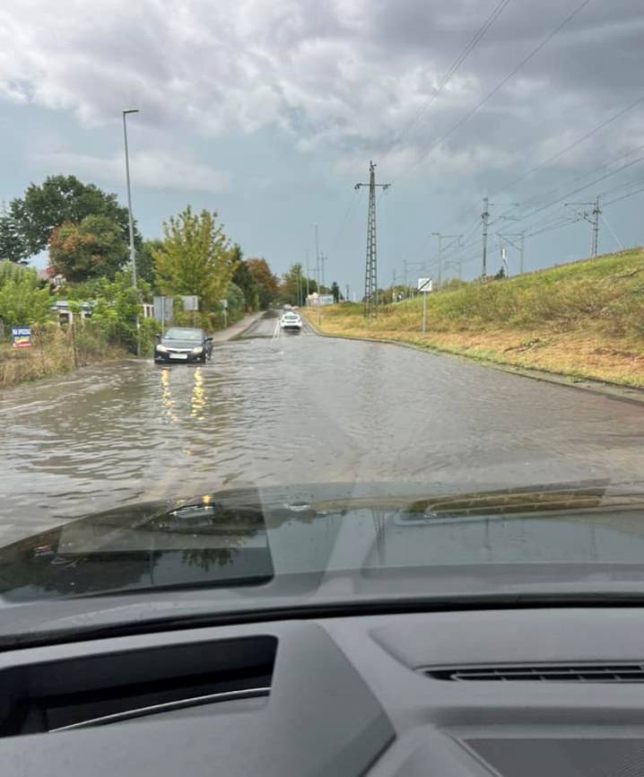 Na dworze, pochmurny dzień. Zdjęcie przez szybę auta na zalaną ulicę. Na przodzie po lewej auto przejedża przez wodę. Naokoło drzewa i słupy z drutami