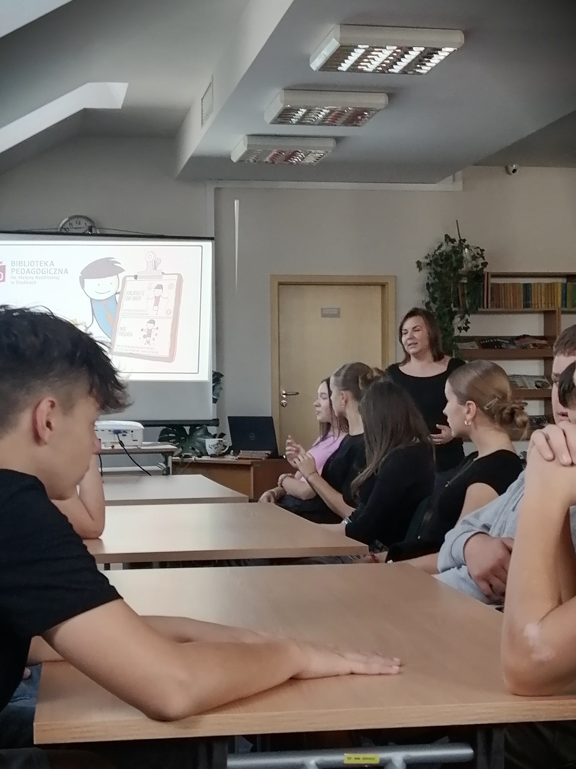 Wnętrze czytelni. Przy stołach siedza nastolatki. Po prawej stoi kobieta