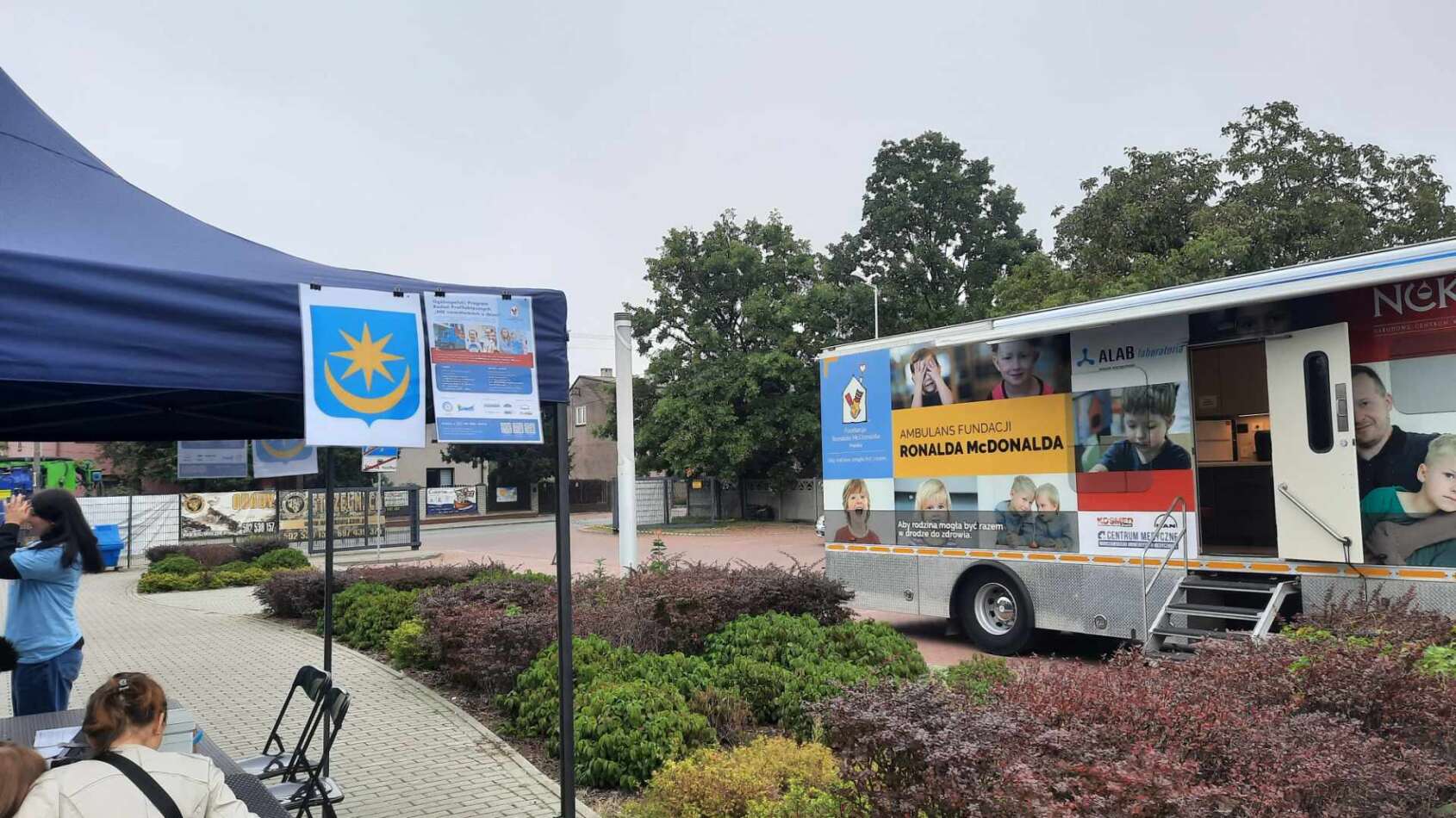 Na dworze, w ciągu dnia. Po lewej widok na niebieski namiot. Po prawej widok na oklejony ambulans