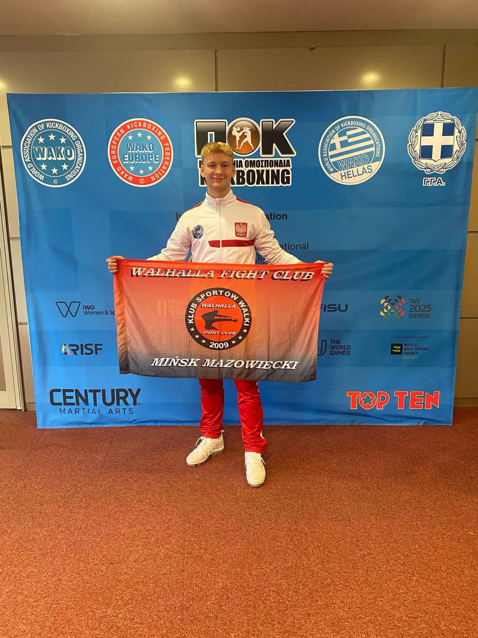 Pomieszczenie. Przed niebieskim banerem usmiechnięty mężczyzna w biało-czerwonym stroju. Trzyma przed sobą czerwoną flagę z napisami klubu sportowego