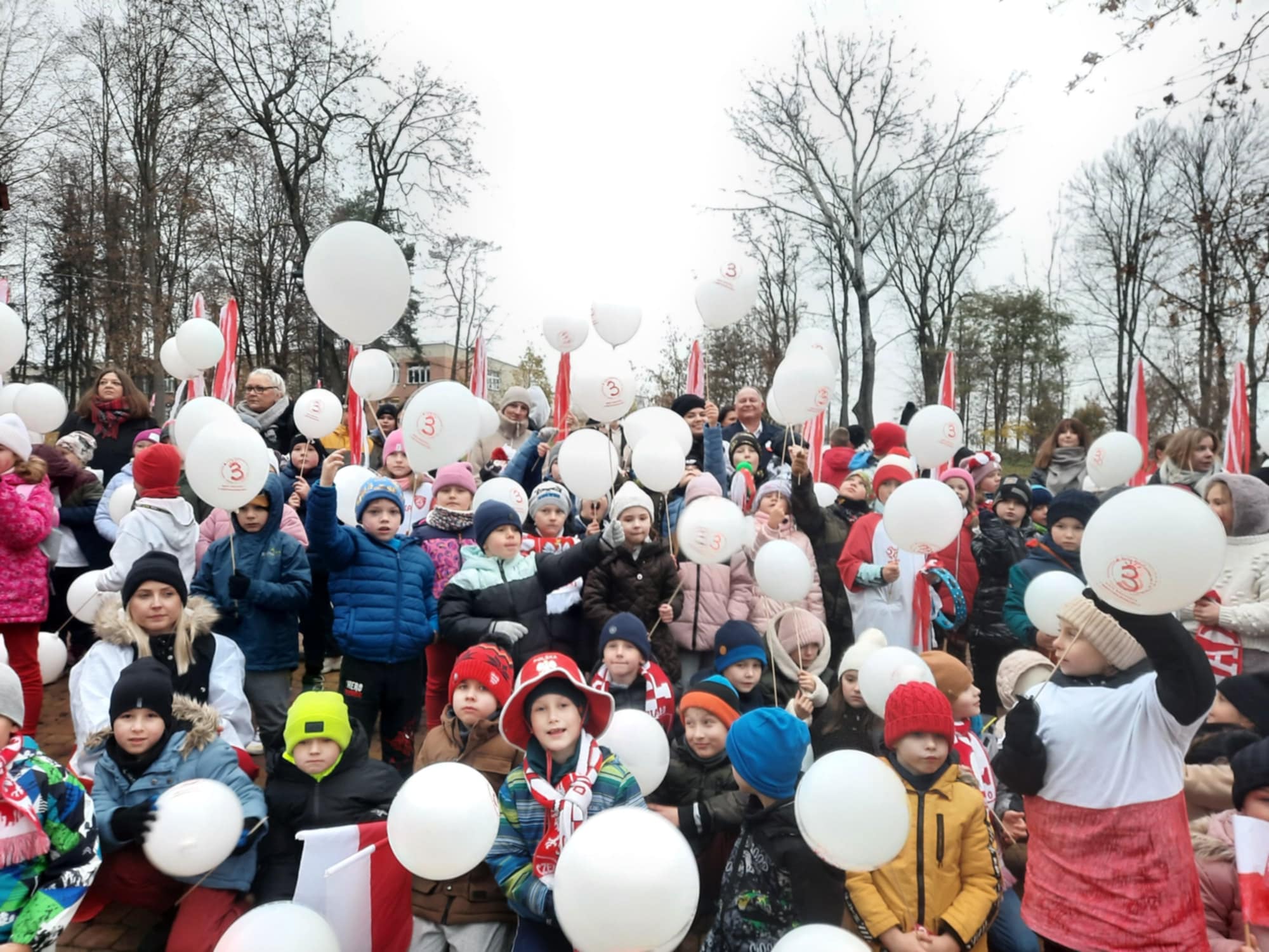 Na dworze w pochmurny dzień. Grupa dzieci w kurtkach i czapkach trzyma białe balony. W tle drzewa bez liści