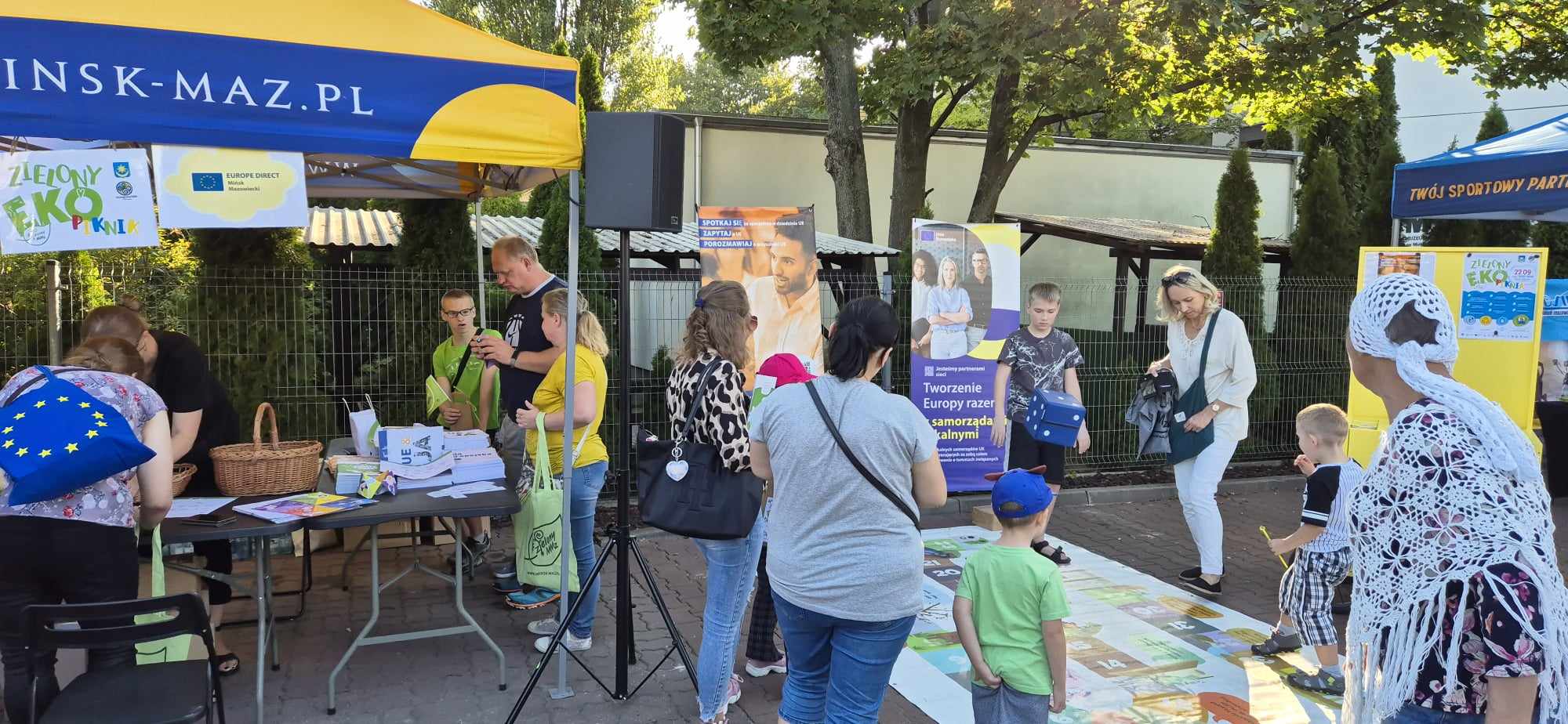 Na dworze, dzień. Grupa dzieci gra w dużą grę planszową rozłożona na podłodze. Naokoło dorośli. Po lewej namiot i obok ludzie