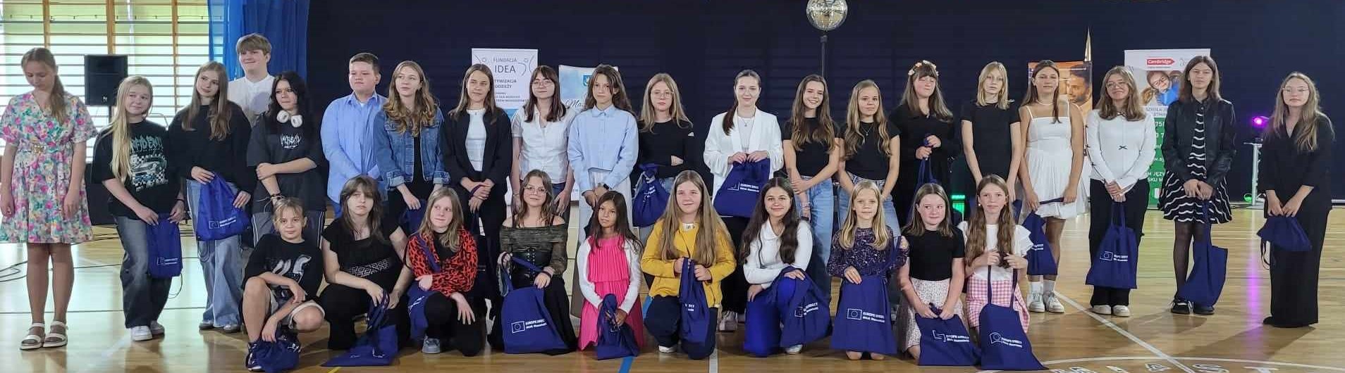 Hala sportowa, dzień. Grupa dzieci z granatowymi torbami. Na przodzie część kuca, reszta w drugiej linii stoi