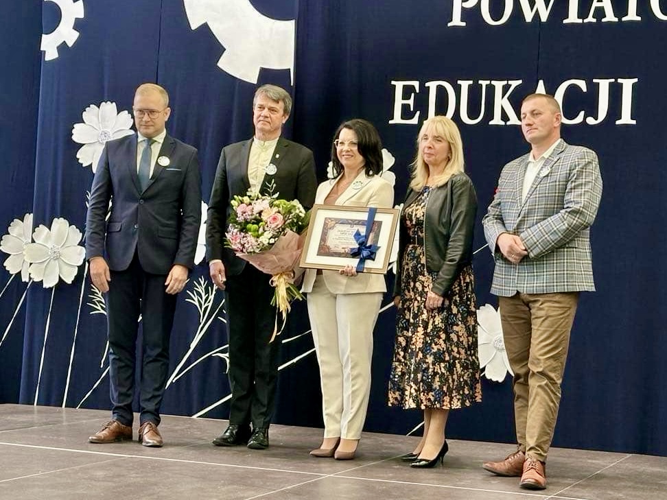 Pomieszczenie. Pięć osób stoi przed granatową kotarą z białymi napisami i kwiatami. Po środku kobieta z bukietem kwiatów i dyplomem. Dyplom w formie obrazka. Ma granatową kokardę