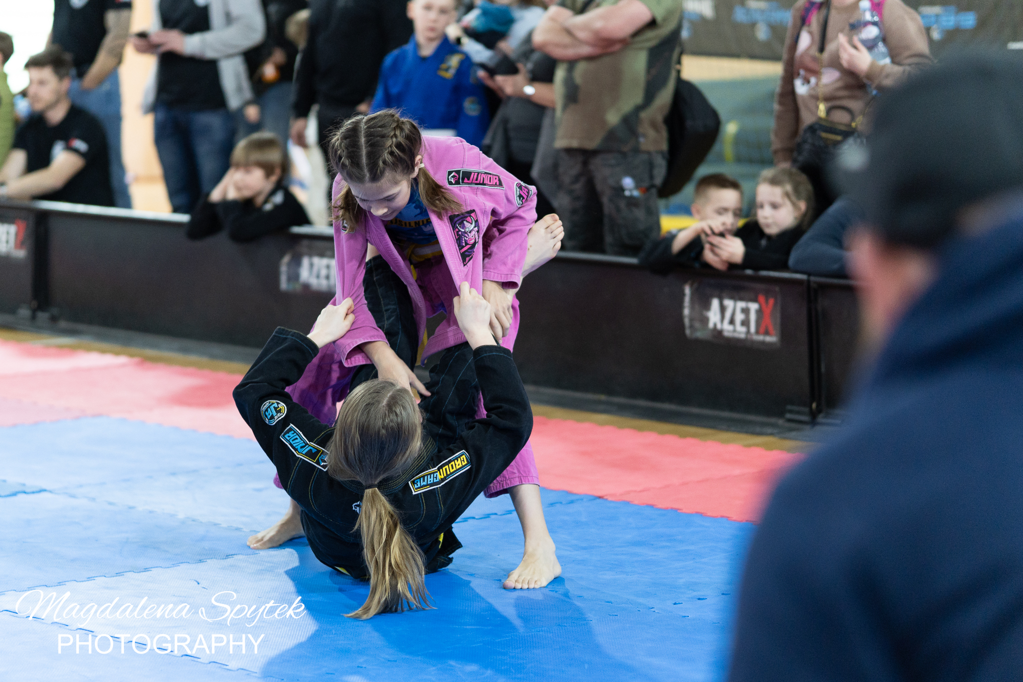 Hala sportowa. Na niebieskich piankowych matach walczą dwie nastolatki. Jedna w czarnym stroju w pozycji połleżącej. Nad nią pochyla się w uchwycie druga w różowym.