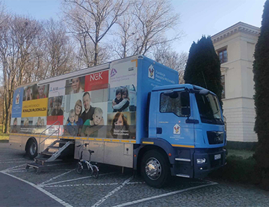 Na placu dzień. Oklejony ambulans. Schodki metalowe do wejścia. W tle drzewa