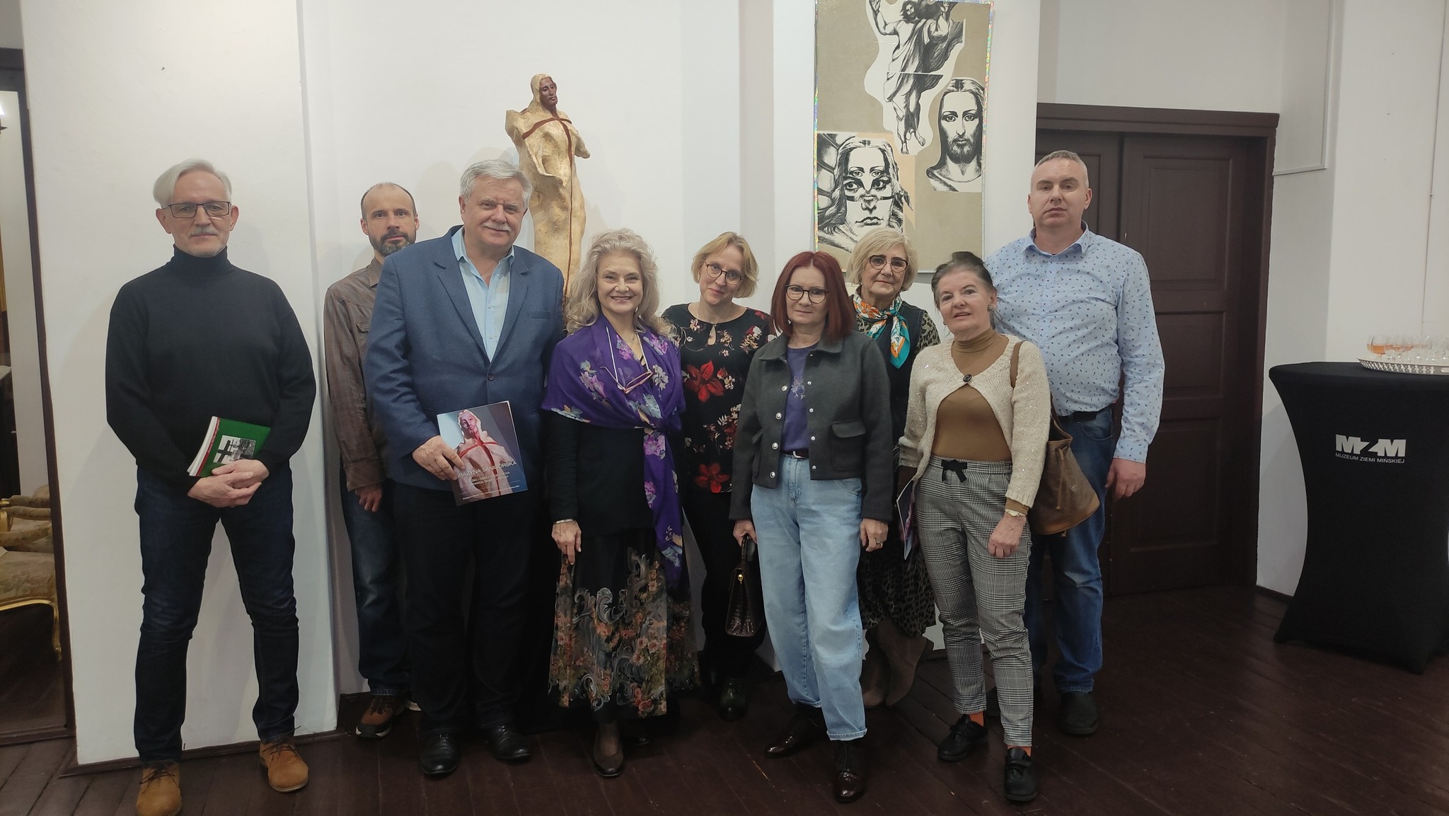 Oświetlone pomieszczenie. Grupa osób pozuje. W tle biała ścian az rzeźbą