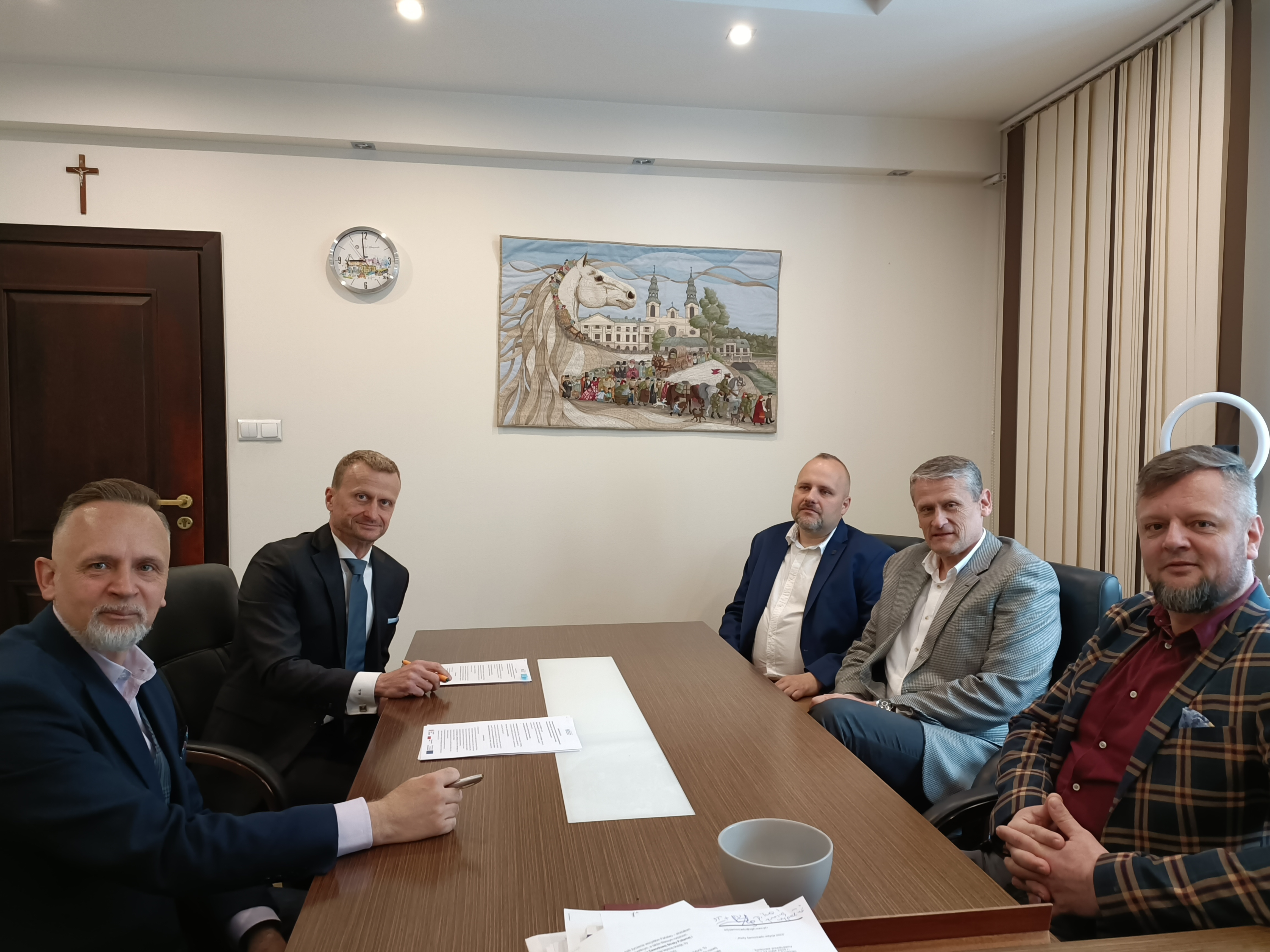 Jasne pomieszczenie. Pięciu mężczyzn siedzi przy stole. Na stole dwa egzemplarze umowy. Na przodzie na jasnej ścianie obraz z koniem i po lewej zegar