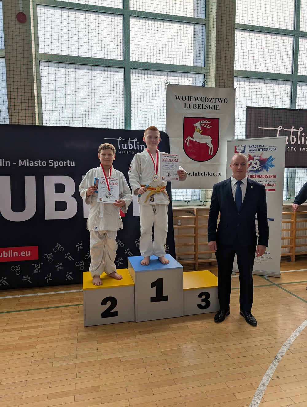 Jasna naturalnym światłem hala. Na parkiecie podium. Na nim dwóch zawodników. Po prawej stronie podium mężczyna w garniturze.
