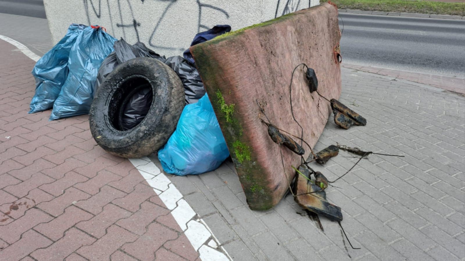 Dzień. Na chodniku oparte o beton stara opona, materac oraz worki