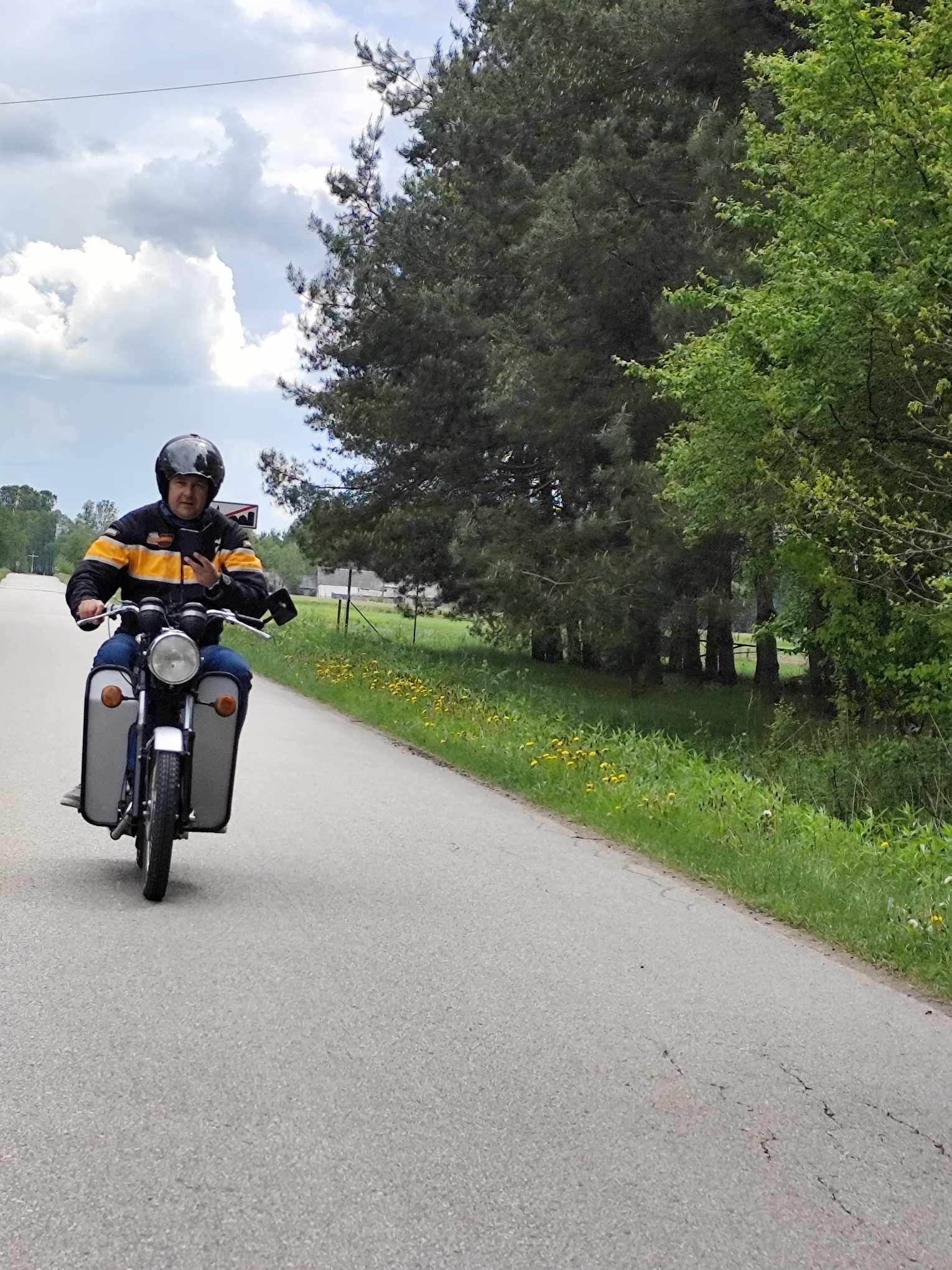 Na dworze, dzień. Motocyklista w kasku jedzie motoem po ulicy. Po prawej zielone drzewa i trawa