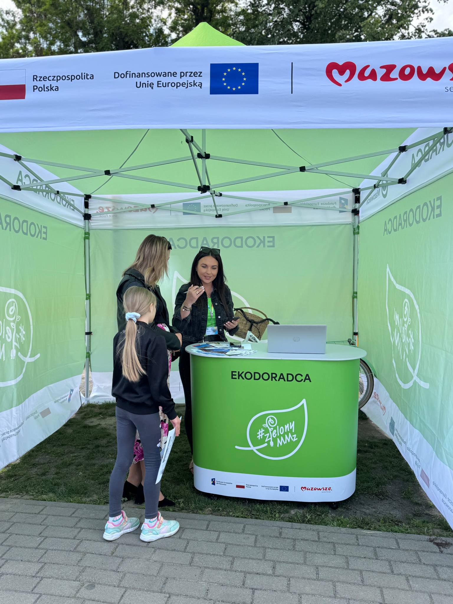 Na dworze, dzień. Pod zielonym namiotem kobieta przy zielonej trybunce. Obok kobieta z dziewcznką słucha