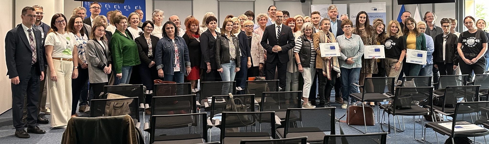 Wnętrze budynku. Duża grupa ludzi pozuje do zdjęcia, przed nimi są krzesła.