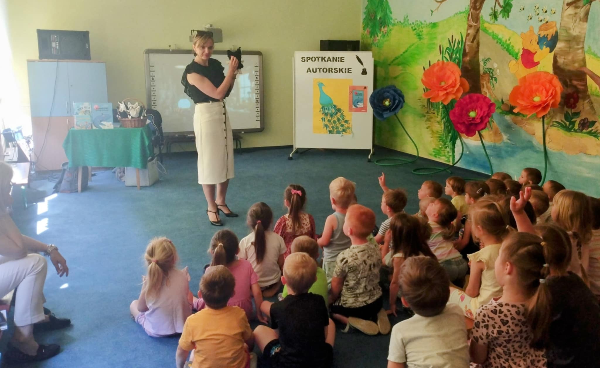 Oświetlona sala przedszkolna. Na niebieskim dywanie tyłem siedzi grupa dzieci. Na przodzie przed dzieśmi stoi kobieta