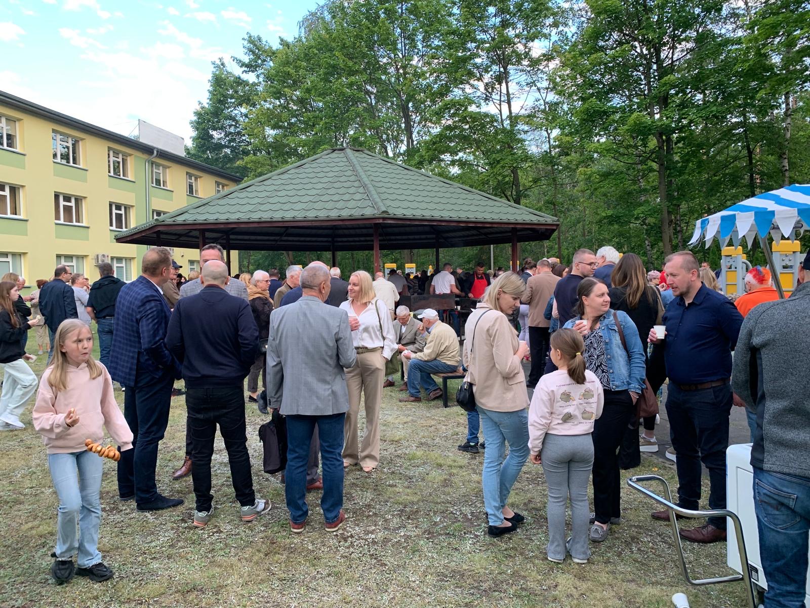 Na dworze. Pochmurny dzień. Spotkanie grupy osób. Część osób trzyma jedzenie i napoje. Po środku altana z zielonym dachem