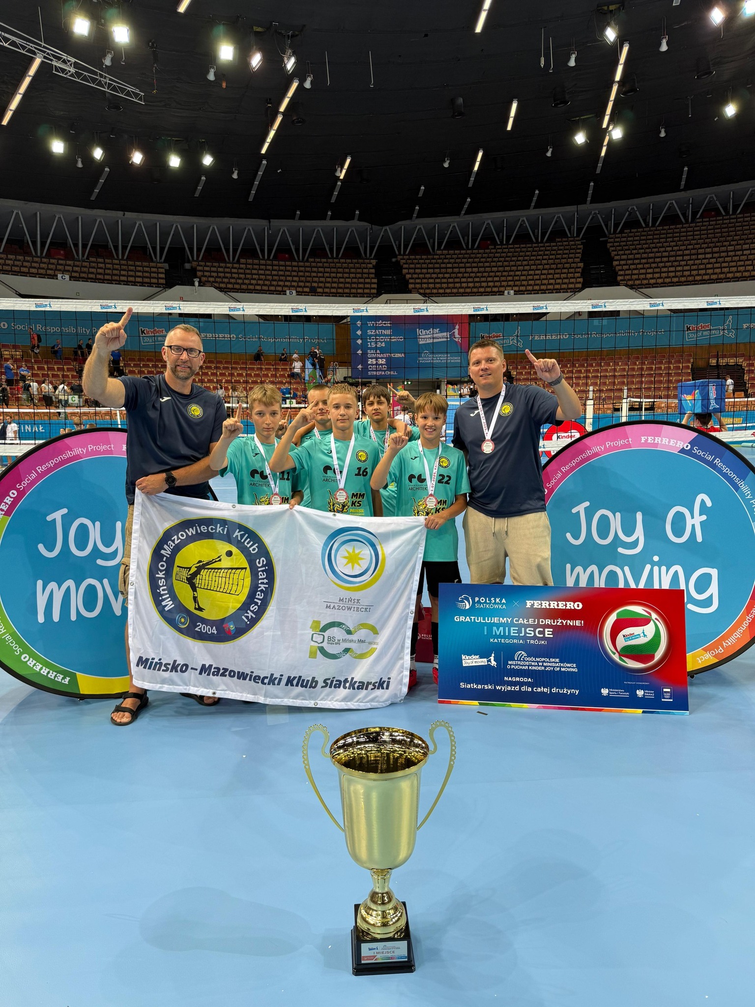 Drużyna pozuje z wielkim pucharem, medalami i banerem zwycięzców na tle hali Spodek. Po obu stronach chłopców trenerzy