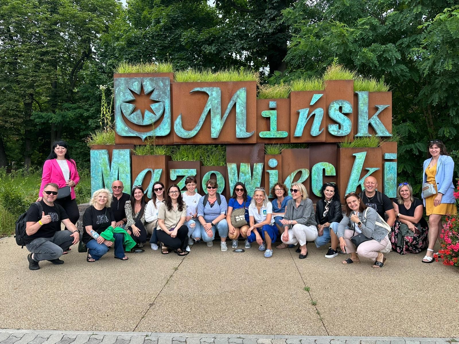 Dzień. Przed napisem Mińsk Mazowiecki grupa osób. W tle drzewa