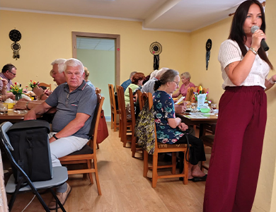 Oświetlone pomieszczenie. Na przodzie kobieta w borodwych spodniach przemawia do mikrofonu, za nią po obu stronach stoł, naokoło których siedzą seniorzy