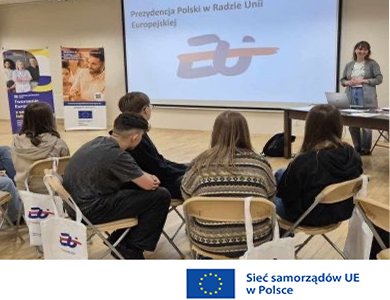 Oświetlone pomieszczenie. Grupa osób słucha kobiety, siedząc na krzesłach. Na przodzie na ekranie treść. Na dole na jasnym logo unii