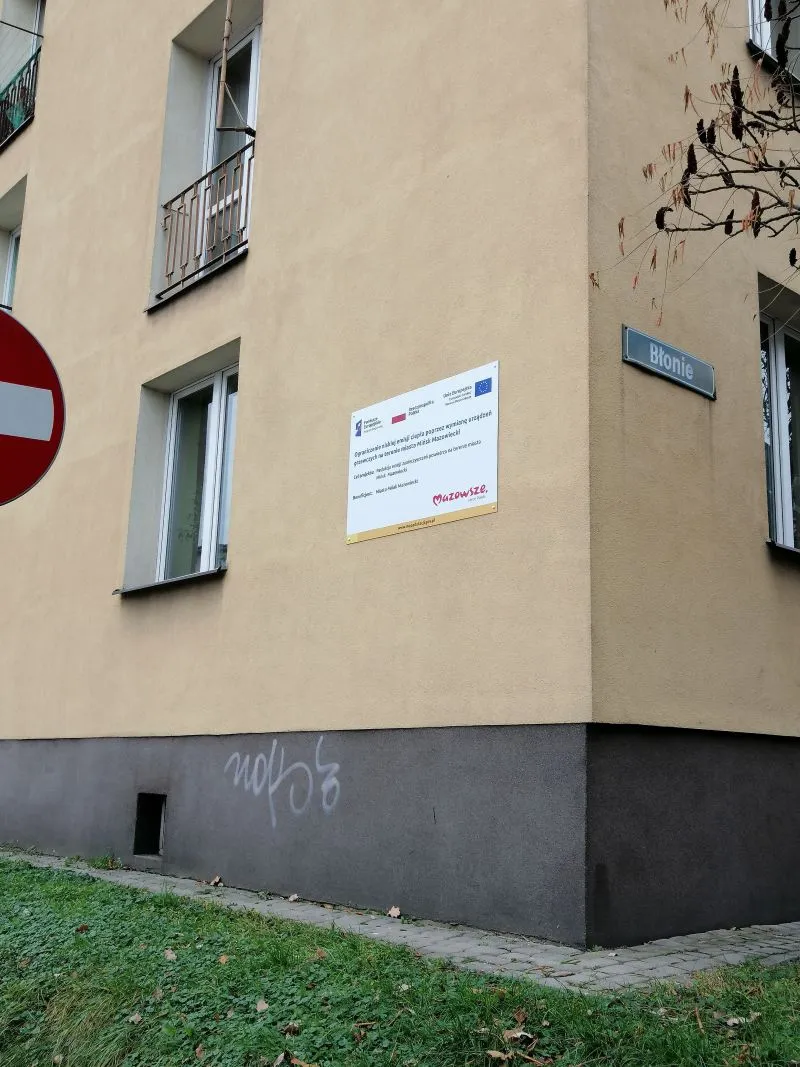 Żółty budynek z tablicą informacyjną, znakiem drogowym zakazu wjazdu i tabliczką uliczną o nazwie Błonie.