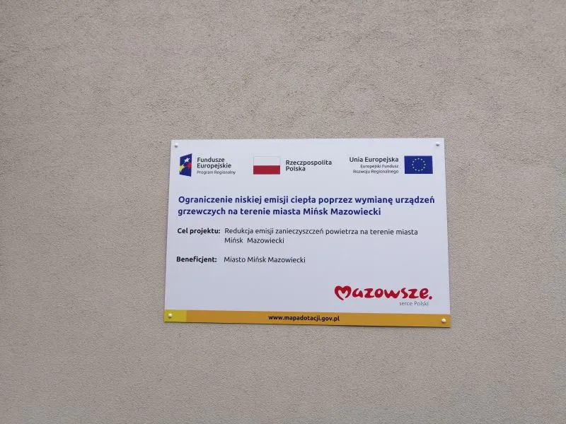 Tablica informacyjna o projekcie ograniczenia niskiej emisji ciepła poprzez wymianę urządzeń grzewczych na terenie miasta Mińsk Mazowiecki, z logotypami Funduszy Europejskich, Rzeczpospolitej Polski i Unii Europejskiej.