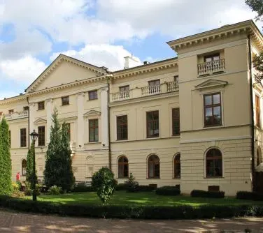 Duży, jasnobeżowy budynek z klasycznymi elementami architektonicznymi otoczony zielonymi drzewami i krzewami.