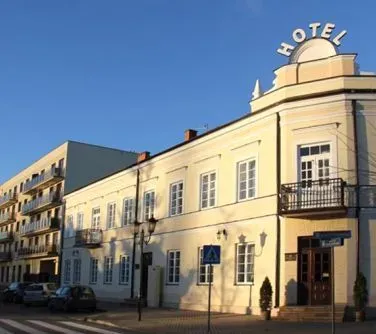 Budynek hotelu w jasnym kolorze z podświetlanym napisem "HOTEL" na dachu i balkonem przy wejściu.