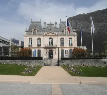 Klasycystyczny budynek z trzema flagami przed wejściem, otoczony ogrodzeniem i położony u podnóża skalistego wzgórza.