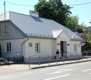 Biały budynek z dwuspadowym dachem pokrytym blachą, z kilkoma oknami i wejściem, przed którym stoją dwie osoby.