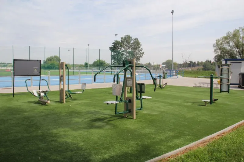 Plenerowa siłownia z różnymi urządzeniami do ćwiczeń na sztucznej trawie i ogrodzona niebieska bieżnia lekkoatletyczna w tle.