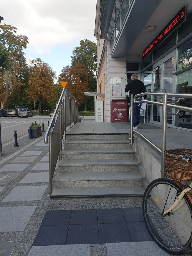 Schody z poręczami prowadzące do wejścia do budynku z otwartymi drzwiami, obok stoi mężczyzna, a po prawej stronie zaparkowany jest rower z wiklinowym koszykiem.