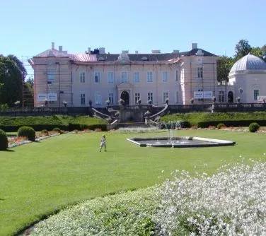 Duży pałac z różowawymi elewacjami i fontanną na zadbanym trawniku z kwiatami.