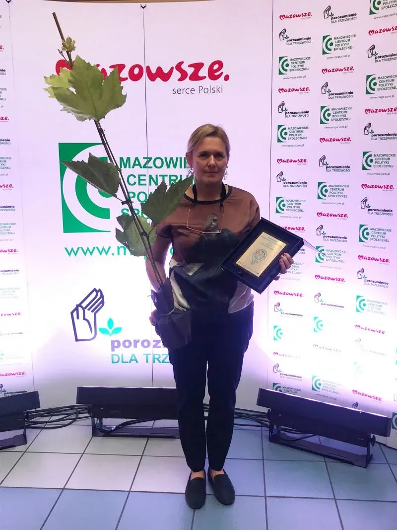 Kobieta stoi trzymając dużą gałąź z liśćmi w jednej ręce oraz ramkę z dyplomem w drugiej na tle ściany z logo Mazowsze i Mazowieckiego Centrum Polityki Społecznej.